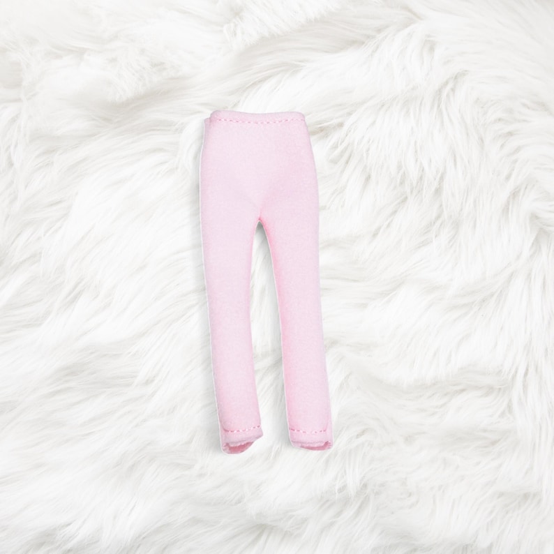 May include: A pair of light pink doll pants featuring a simple, fitted design with a high waist and straight legs. The garment is constructed from a solid, smooth fabric with visible stitching along the hem and inner seams of the pant legs.