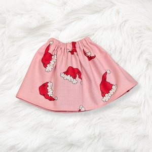 May include: A light pink skirt featuring an all-over print of red Santa hats with white trim. The skirt has an elasticized waistband and a flared silhouette, showcasing a repeating pattern of festive holiday headwear across the fabric.