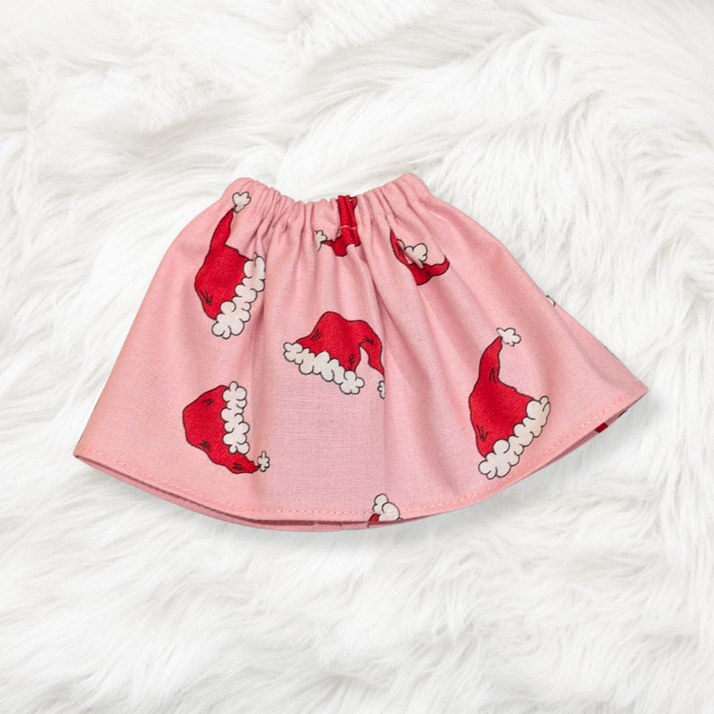 May include: A light pink skirt featuring an all-over print of red Santa hats with white trim. The skirt has an elasticized waistband and a flared silhouette, showcasing a repeating pattern of festive holiday headwear across the fabric.