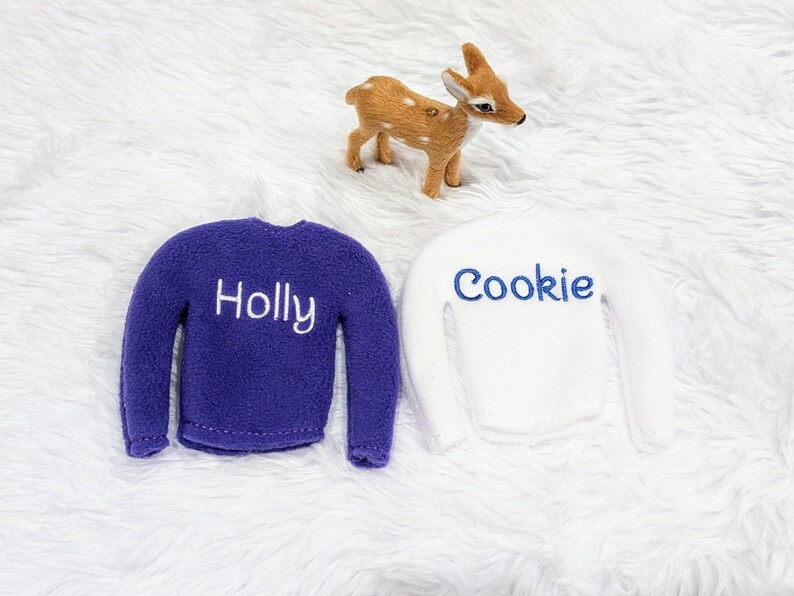 May include: Two elf-sized sweaters, one purple with "Holly" embroidered in white and one white with "Cookie" embroidered in blue.