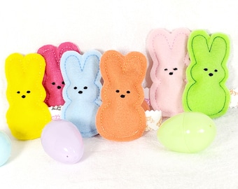 May include: A collection of six felt bunny-shaped ornaments in yellow, hot pink, light blue, orange, pale pink, and lime green, each featuring stitched outlines and embroidered facial details, accompanied by three plastic Easter eggs in pastel colors.
