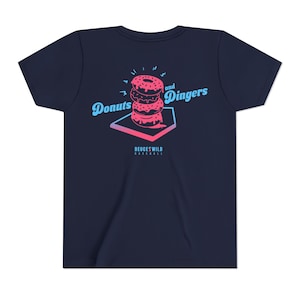 May include: Navy blue t-shirt featuring a stacked donut graphic with the words "Donuts and Dingers" in a stylized font. The graphic includes a baseball home plate design and the text "DEUCESWILD BASEBALL".