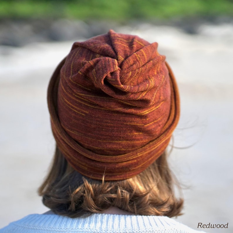 Pu&ograve; includere: Un cappello in stile turbante color ruggine con strisce gialle. Il cappello &egrave; realizzato in un tessuto morbido e strutturato e viene indossato sulla testa. La parola "Redwood" &egrave; stampata nell'angolo in basso a destra dell'immagine.
