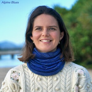 Pu&ograve; includere: Una donna indossa un scaldacollo blu e un maglione color crema a trecce con ricami floreali. Il testo "Alpine Blues" &egrave; nell'angolo in alto a sinistra. Lo sfondo &egrave; una scena di spiaggia con un ponte e alberi.
