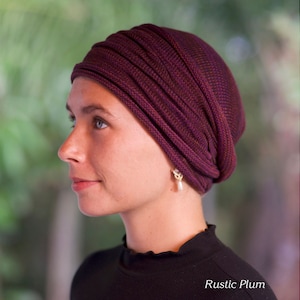 Puede incluir: Una mujer lleva un turbante de punto morado y marr&oacute;n con un pendiente de perla. El texto "Rustic Plum" es visible en la parte inferior de la imagen.