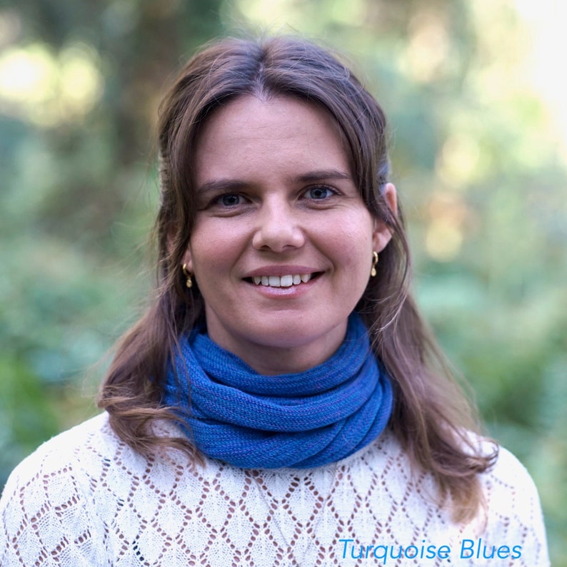 Op de afbeelding: Een vrouw met lang bruin haar draagt een witte trui met een blauwe sjaal. Ze glimlacht en kijkt naar de camera. De achtergrond is wazig en onscherp.