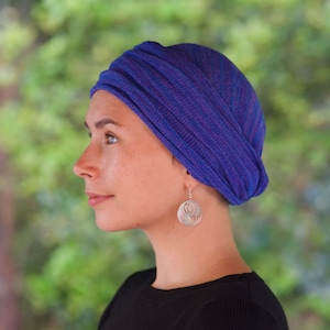 Op de afbeelding: Een vrouw draagt een blauwe en roze gestreepte turban met een zilveren ronde oorbel. De turban is om haar hoofd gewikkeld en achterop vastgebonden.