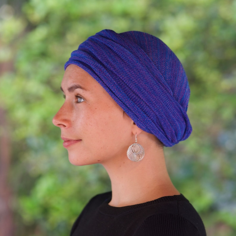 Op de afbeelding: Een vrouw draagt een blauwe en roze gestreepte turban met een zilveren ronde oorbel. De turban is om haar hoofd gewikkeld en achterop vastgebonden.