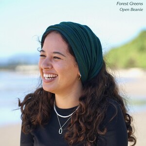 Pu&ograve; includere: Una donna indossa un berretto aperto verde scuro. Il berretto &egrave; realizzato in un morbido tessuto a coste. Il testo "Forest Greens Open Beanie" si trova nell'angolo in alto a destra. Indossa una maglia nera e una collana d'argento.