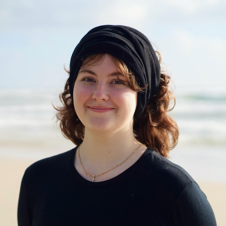 Pu&ograve; includere: Una persona indossa un copricapo nero e una maglietta nera, con una collana d'oro. Lo sfondo &egrave; una spiaggia con l'oceano in lontananza. La persona sorride.
