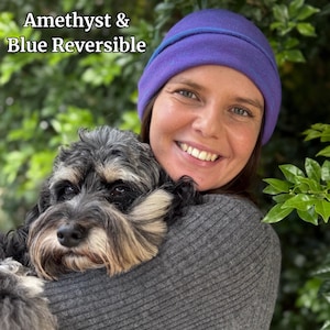 Pu&ograve; includere: Berretto reversibile ametista e blu. Il cappello &egrave; viola con un bordo blu. Il testo "Amethyst & Blue Reversible" si trova nell'angolo in alto a sinistra. Una persona indossa il cappello e tiene un cane.