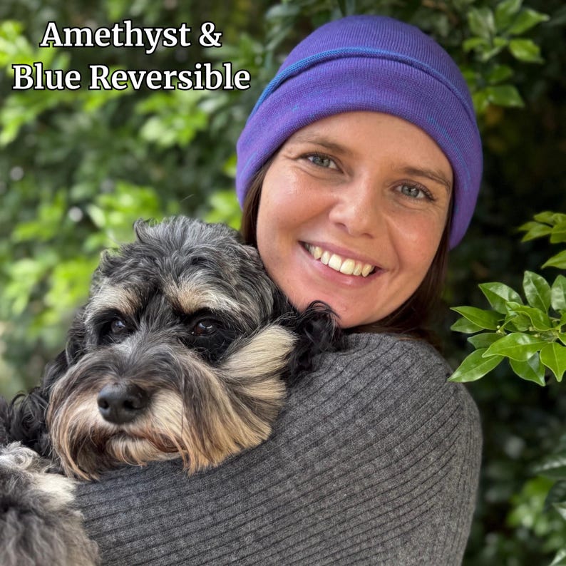 Pu&ograve; includere: Berretto reversibile ametista e blu. Il cappello &egrave; viola con un bordo blu. Il testo "Amethyst & Blue Reversible" si trova nell'angolo in alto a sinistra. Una persona indossa il cappello e tiene un cane.