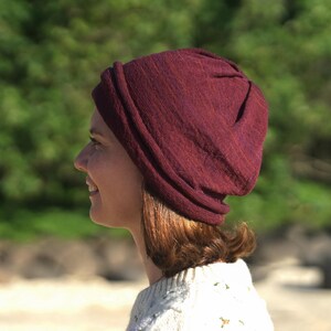 Pu&ograve; includere: Un berretto color bordeaux realizzato in un tessuto morbido e strutturato. Il berretto &egrave; indossato sulla testa e ha una vestibilit&agrave; leggermente ampia. Lo sfondo &egrave; una scena all'aperto sfocata verde e beige.