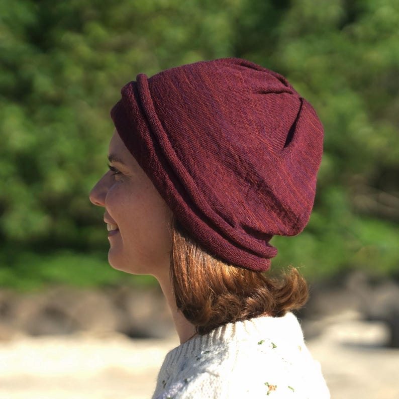 Pu&ograve; includere: Un berretto color bordeaux realizzato in un tessuto morbido e strutturato. Il berretto &egrave; indossato sulla testa e ha una vestibilit&agrave; leggermente ampia. Lo sfondo &egrave; una scena all'aperto sfocata verde e beige.