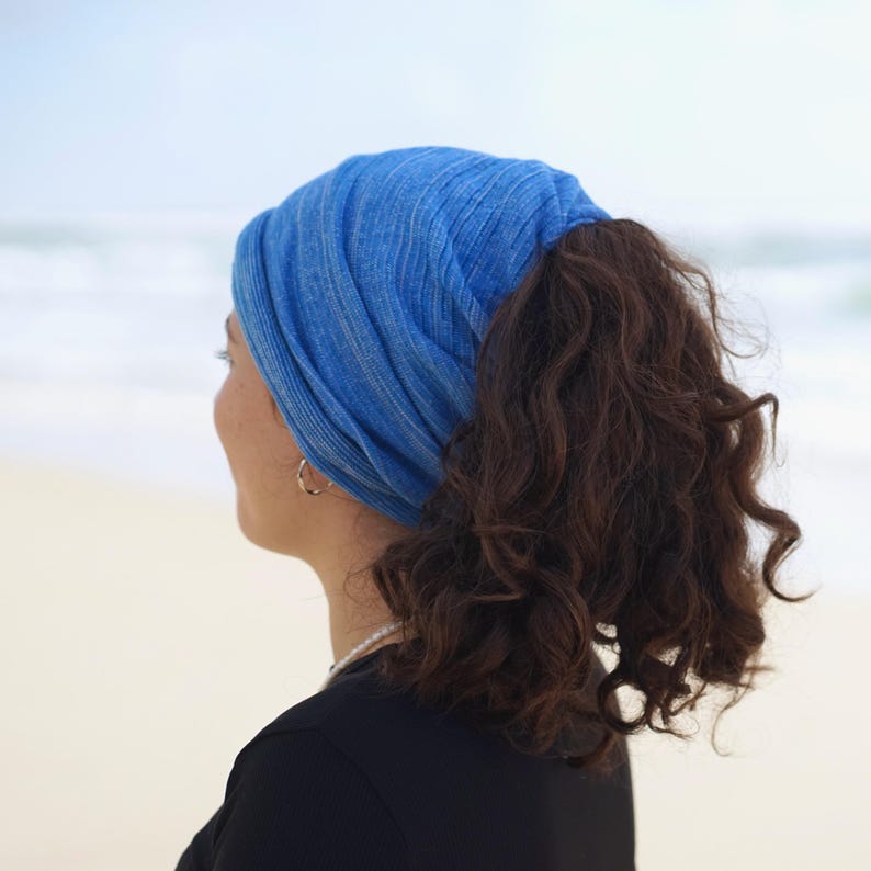 Pu&ograve; includere: Una fascia blu con un'apertura per una coda di cavallo. La fascia &egrave; realizzata in un tessuto morbido e strutturato. La donna indossa una maglietta nera e una collana di perle. Lo sfondo &egrave; una spiaggia con l'oceano in lontananza.