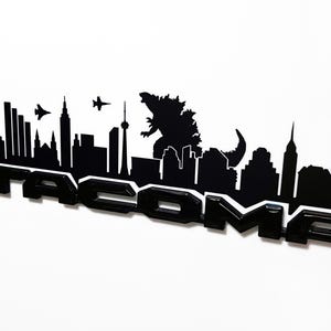 May include: Black Tacoma emblem featuring a city skyline silhouette with Godzilla and two fighter jets. The emblem is glossy black with a white outline, set against a white background. A unique automotive accessory.