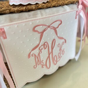 Puede incluir: Una pancarta de tela blanca con lunares rosas y un lazo y un monograma bordados en rosa. La pancarta tiene un borde festoneado y est&aacute; unida a una cesta de mimbre.