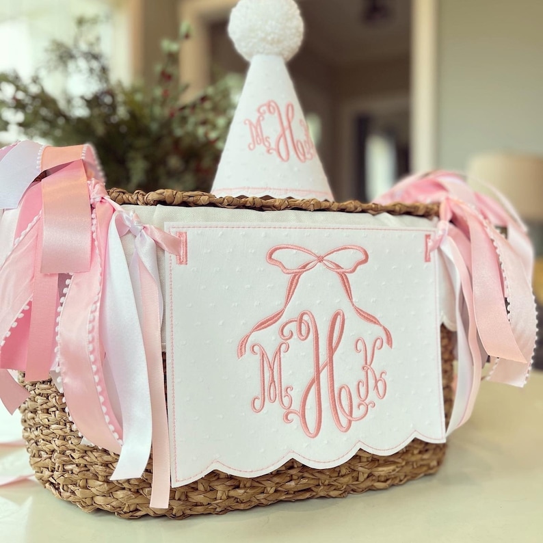 Puede incluir: Una cesta de mimbre blanca con una guirnalda de cintas rosa y blanca y una pancarta blanca con letras bordadas en rosa. La pancarta dice "M.H.K." y tiene un lazo rosa sobre el monograma. Un gorro de fiesta blanco con un pomp&oacute;n rosa y letras bordadas en rosa est&aacute; en la parte superior de la cesta.
