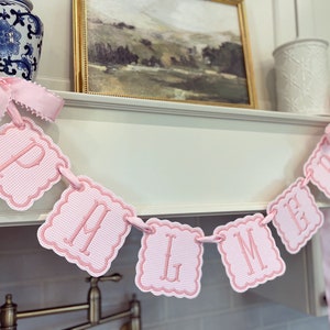 May include: Pink and white banner with the letters "PHMGB" in a cursive font. The banner is made of fabric and has a scalloped edge. It is hanging from a shelf.