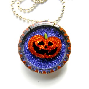 May include: A silver chain necklace with a round pendant featuring a glittery orange pumpkin with a black jack-o'-lantern face on a blue glitter background.