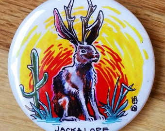 May include: A colourful button featuring a jackalope, a mythical rabbit with antlers, against a sunset backdrop with a cactus and grass. The button reads "JACKALOPE".