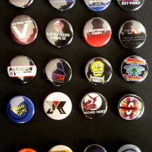 May include: A collection of 16 round, colorful buttons with various video company logos, including Cannon, AETOS, Embassy Home Entertainment, Key Video, VESTRON VIDEO, LIGHTNING VIDEO, TRANS WORLD ENTERTAINMENT, PARAGON, PRISM ENTERTAINMENT, MASSACRE VIDEO, VIDEO GEMS, X, WIZARD VIDEO, R RESTRICTED, PG RATED, and VHS.