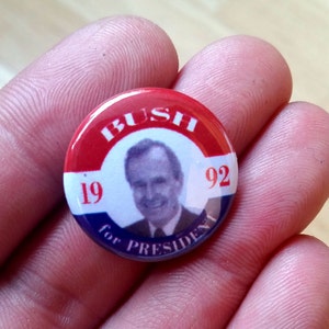 May include: A circular political badge featuring a portrait of George H.W. Bush. The badge has a red and white border with the word "BUSH" at the top and "1992" on the sides. The words "for PRESIDENT" are at the bottom.