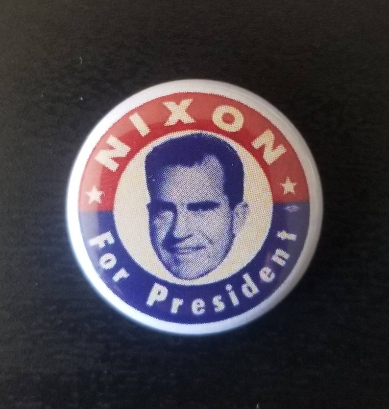 May include: A round button with a portrait of Richard Nixon, with the text "NIXON For President". The button has a red, white, and blue color scheme, with a star on each side of the word "NIXON".