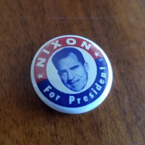 May include: A vintage campaign button with a portrait of Richard Nixon. The button has a white background with red and blue text that reads "NIXON For President". The image of Nixon is in a blue circle.