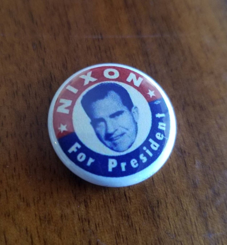 May include: A vintage campaign button with a portrait of Richard Nixon. The button has a white background with red and blue text that reads "NIXON For President". The image of Nixon is in a blue circle.
