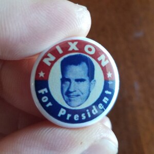 May include: A vintage campaign button with a portrait of Richard Nixon. The button features a red and blue color scheme with the words "NIXON" and "For President" encircling the portrait. A star is visible on the button.