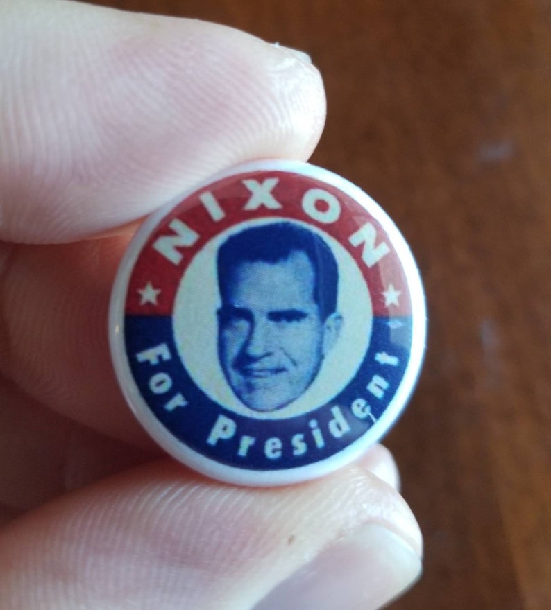 May include: A vintage campaign button with a portrait of Richard Nixon. The button features a red and blue color scheme with the words "NIXON" and "For President" encircling the portrait. A star is visible on the button.