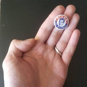 May include: A vintage political campaign button with the text "NIXON For President" encircling a portrait of Richard Nixon. The button has a red, white, and blue color scheme, and is held in a person's hand.