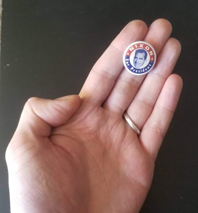 May include: A vintage political campaign button with the text "NIXON For President" encircling a portrait of Richard Nixon. The button has a red, white, and blue color scheme, and is held in a person's hand.
