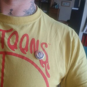 May include: A yellow t-shirt with the word "Toons" in red lettering. A round button with the text "Nixon for President" is pinned on the shirt. The shirt is made of a soft material.
