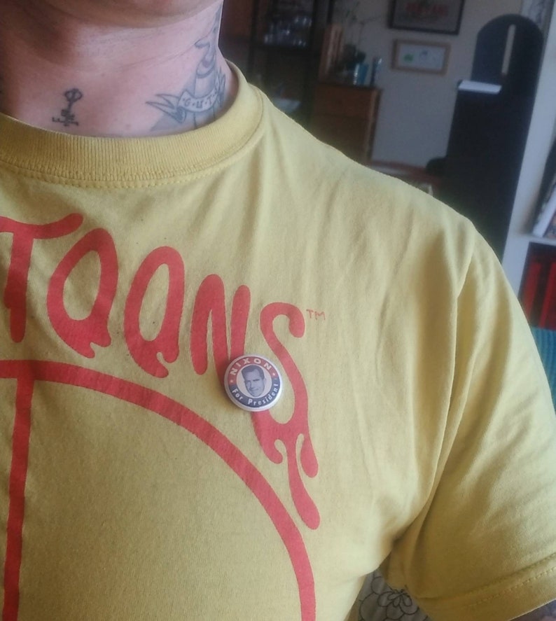 May include: A yellow t-shirt with the word "Toons" in red lettering. A round button with the text "Nixon for President" is pinned on the shirt. The shirt is made of a soft material.