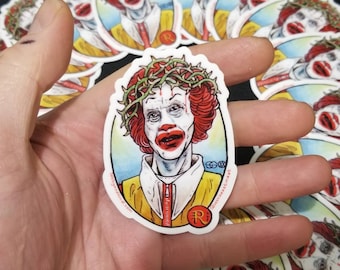 May include: A sticker featuring a cartoon illustration of Ronald McDonald wearing a crown of thorns. The background is a yellow and blue gradient. The sticker is oval shaped and has a red "R" in the bottom right corner.