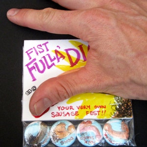 May include: A clear plastic bag containing four colourful button badges. The badges feature illustrations of sausages with the words "Fork Sword", "Dog", "Steak", and "Wank" written on them. The bag also includes a card with the words "Fist Fulla Di*k Your Very Own Sausage Fest!!" written on it.