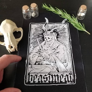 Puede incluir: Parche bordado en blanco y negro que presenta una figura esquelética con cuernos y una corona de espinas, sosteniendo una daga y un cáliz. La figura está sentada en una mesa con un diseño de pentagrama. El texto "blasfemia" está escrito en una fuente gótica debajo de la figura.