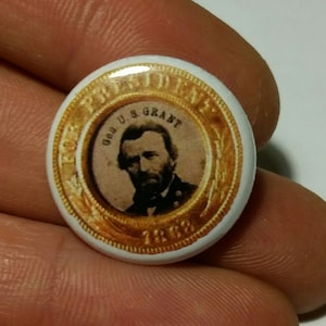 Puede incluir: Un botón blanco con un borde dorado y un retrato de Ulysses S. Grant. El texto "FOR PRESIDENT" y "U.S. GRANT" están escritos alrededor del retrato. El año "1868" está escrito debajo del retrato.