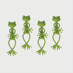 May include: Four green frog-shaped drink markers. Each marker has a frog head, arms, and legs, with a loop at the bottom. The markers are made of a flexible material and are designed to wrap around the stem of a glass.