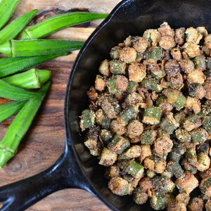 May include: A cast iron skillet filled with crispy fried okra. The okra is a vibrant green color and is coated in a light brown breading. Fresh okra pods are visible in the background.
