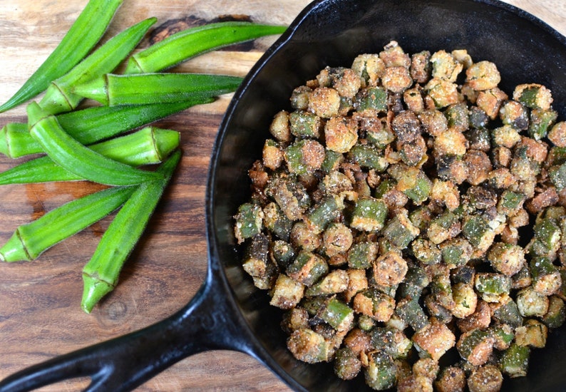 May include: A cast iron skillet filled with crispy fried okra. The okra is a vibrant green color and is coated in a light brown breading. Fresh okra pods are visible in the background.