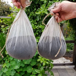 May include: Two white mesh bags filled with dark material, possibly soil or compost, are held up against a backdrop of green foliage. Each bag is tied with twine at the top, creating a rustic, natural aesthetic.