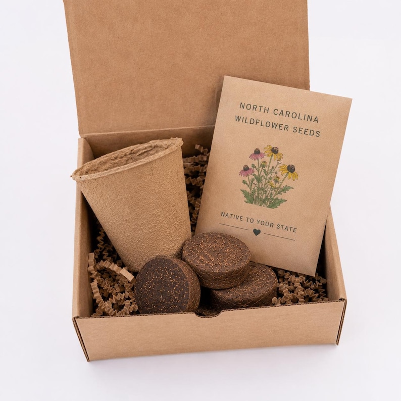 May include: A brown cardboard box containing a tan fiber planting pot, three dark brown soil discs, and a tan paper packet labeled North Carolina Wildflower Seeds Native to Your State with a colorful illustration of yellow and pink flowers.