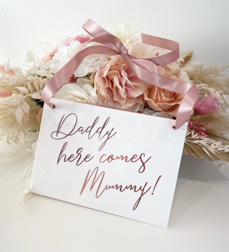 May include: White sign with the text "Daddy here comes Mummy!" in rose gold lettering. A pink satin ribbon is tied around the sign.