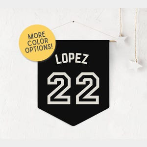 May include: Black pennant banner with the name "LOPEZ" and the number "22" in white block letters. A yellow circle with the text "MORE COLOR OPTIONS!" is in the upper left corner. The banner is hanging from a wooden dowel.