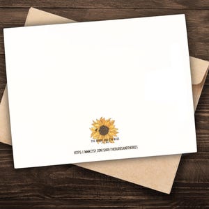 May include: A white greeting card with a sunflower illustration and a kraft paper envelope. The card features a watercolor sunflower design with the text "The Burrs and the Bees". The card is on a dark wood surface.
