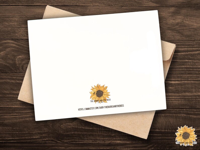 May include: A white greeting card with a sunflower illustration and a kraft paper envelope. The card features a watercolor sunflower design with the text "The Burrs and the Bees". The card is on a dark wood surface.