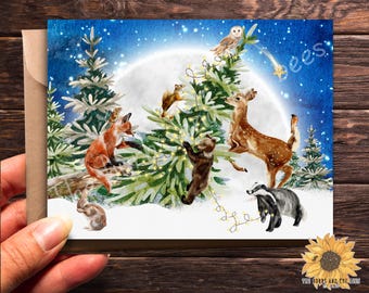 May include: A watercolour illustration of woodland animals decorating a Christmas tree. A fox, deer, bear, squirrel and badger are gathered around the tree, which is decorated with twinkling lights. The background is a starry night sky with a full moon.